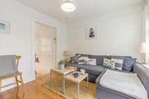 A modern living room with a grey sofa, a wooden chair, a small table with a plant and a mug, and a door leading to another room.