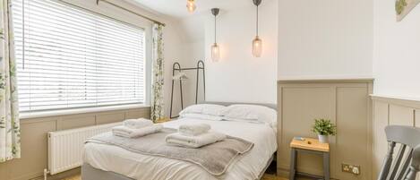 Un dormitorio con cama, mesita de noche, silla, radiador, ventana con cortinas e iluminación en la pared.