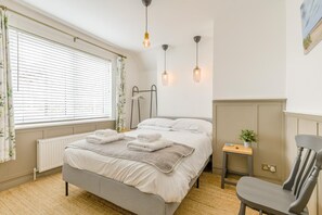 A bedroom with a bed, bedside table, chair, radiator, window with curtains, and wall-mounted lights.