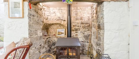A stone-walled room with a wood-burning stove, a framed picture, and a wicker basket with firewood.