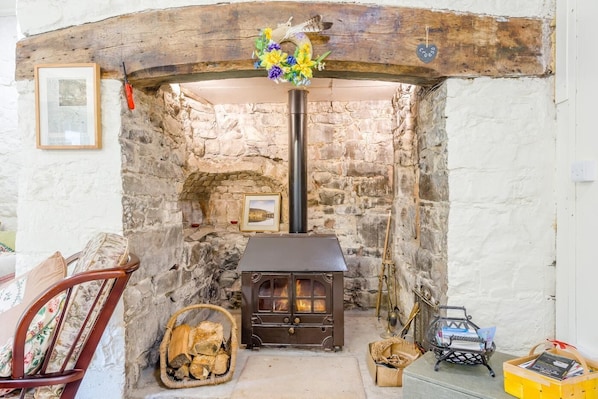 A stone-walled room with a wood-burning stove, a framed picture, and a wicker basket with firewood.