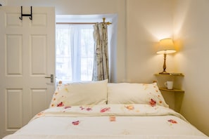 A bed with floral bedding, a bedside lamp, a cup on a nightstand, and a window with curtains.
