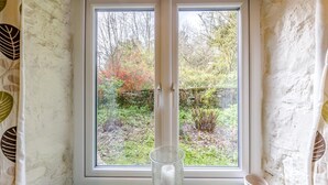 Una habitación con ventana que da a un jardín, un florero de vidrio transparente en el alféizar y un cuenco blanco a la derecha.