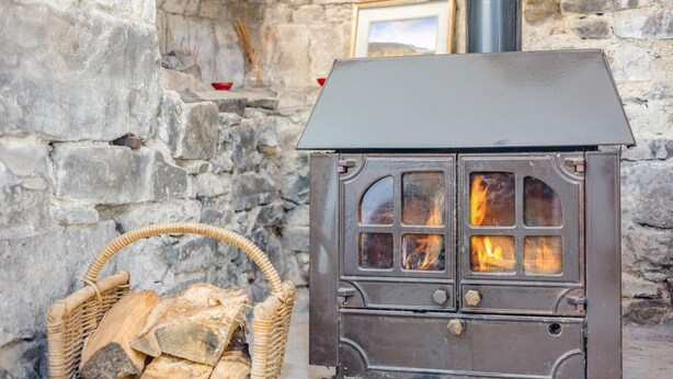 Una estufa de leña con chimenea, una pared de piedra, un cuadro enmarcado y una cesta de mimbre con leña.