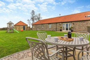 A patio with a table set for breakfast, a stone building with a red-tiled roof, and a children's play structure. - 3 Bed in nr Beadnell (oc-cr1279) (Beadnell)
