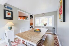 Un coin repas avec une table en bois, des chaises blanches, un panier de fruits et une lampe posée sur une petite commode.