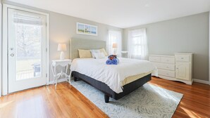 Une chambre à coucher avec un lit, des tables de chevet, une commode et une vue sur l’extérieur, visible à travers une porte.