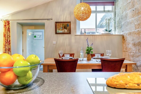 Un comedor con una mesa dispuesta para dos personas, un cuenco de fruta sobre la mesa y una hogaza de pan en la encimera.