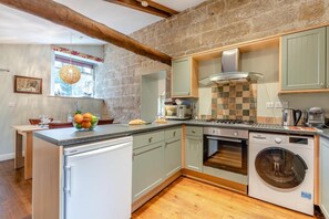 A compact kitchen with a built-in oven, microwave, and washing machine, featuring a stone wall and wooden beams. - 2 Bed in Fylingdales (oc-wa313) (Fylingdales)