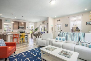 A spacious living room with a white sofa, a red armchair, and a coffee table. A kitchen with wooden cabinets and a dining area with a table and chairs are visible in the background. - Welcome to Slice of Haven with HOT TUB! (South Haven)