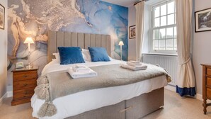 Ett sovrum med en stor säng, två sänglampor, ett träbord vid sängen, ett stort fönster med gardiner och en väggmålning.