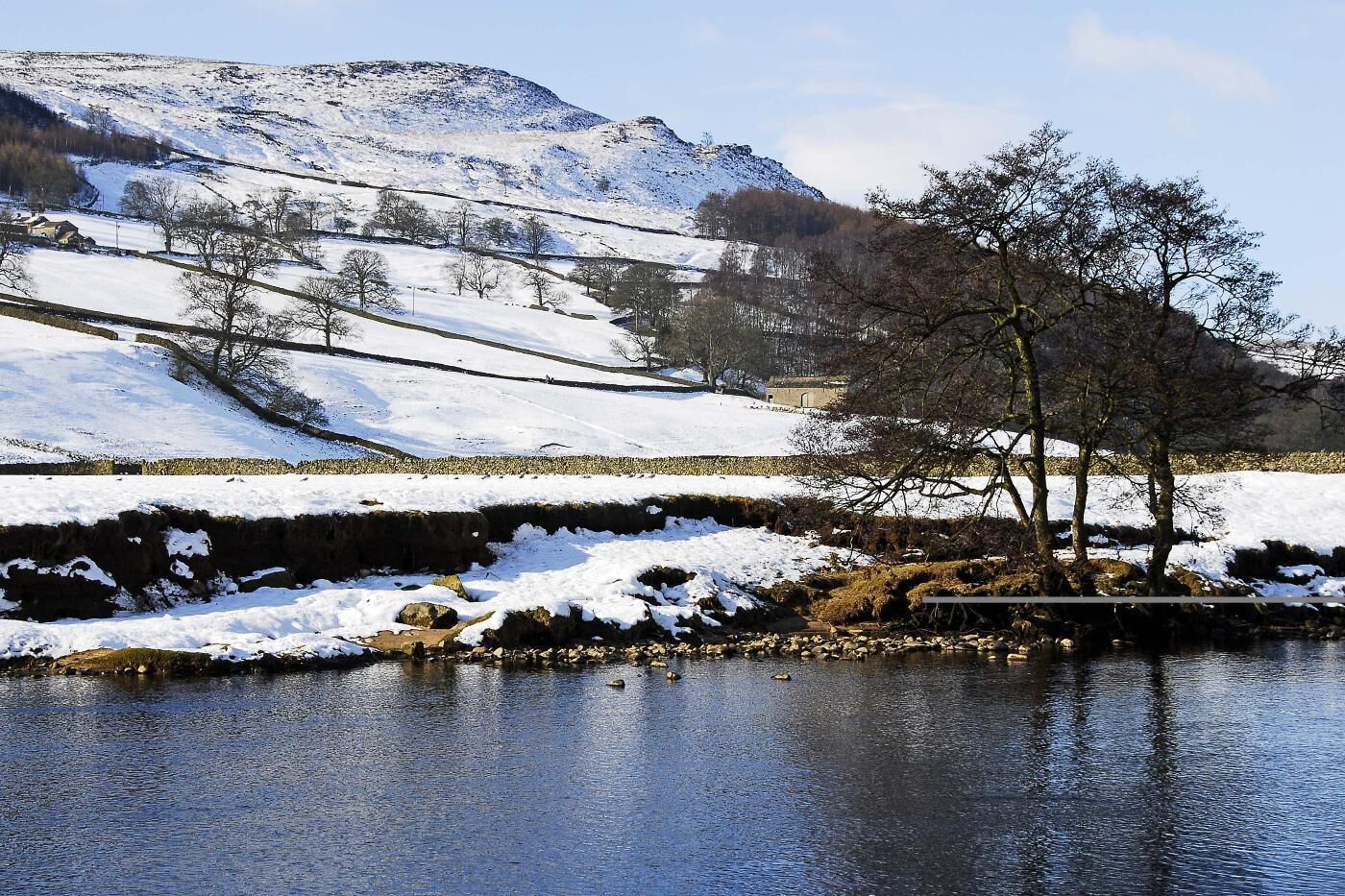 A snow-covered landscape with a body of water, trees, and a distant hill.