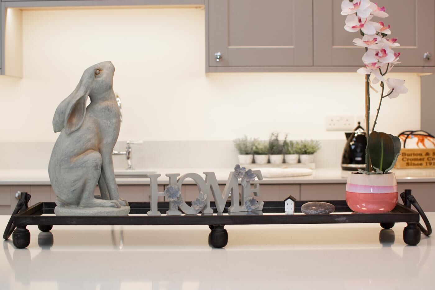 Un comptoir de cuisine orné d’une sculpture de grenouille en pierre, d’une enseigne « HOME », d’une petite maquette de maison et d’une orchidée en pot.