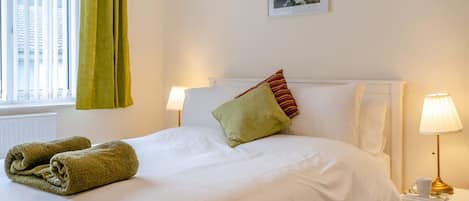 Cama bem arrumada com lençóis brancos, um travesseiro verde e toalhas dobradas ao lado. Um criado-mudo com luminária e uma xícara. Uma moldura com um quadro na parede acima da cama.