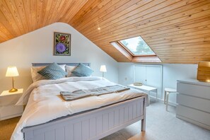 A bedroom with a sloped ceiling, a bed with white bedding, two bedside lamps, a small table, and a dresser.