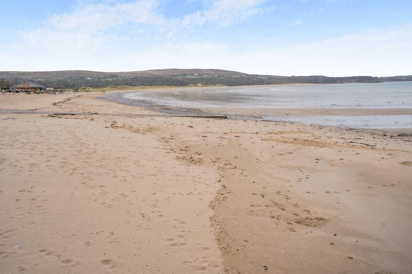 Ein Sandstrand mit Fußspuren, ein ruhiges Meer und sanfte Hügel in der Ferne.