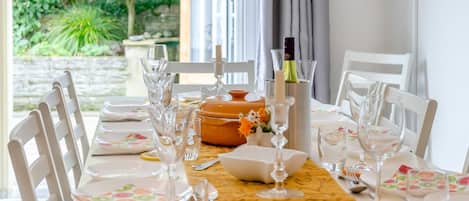 A dining table set with white chairs, plates, and glassware, with a view of greenery outside.