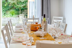 A dining table set with white chairs, plates, and glassware, with a view of greenery outside. - 5 Bed in Oxwich (oc-g31916) (Oxwich)
