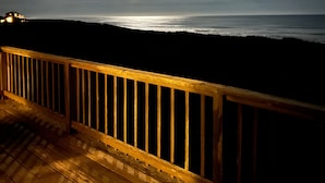 Eine Holzterrasse mit Geländer blickt auf einen Strand in der Nacht, unter einem teilweise bewölkten Himmel mit sichtbar erkennbarem Mond.