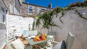 Une terrasse avec une table mise pour le petit-déjeuner, comprenant des croissants et des fruits, entourée de chaises blanches agrémentées de coussins.