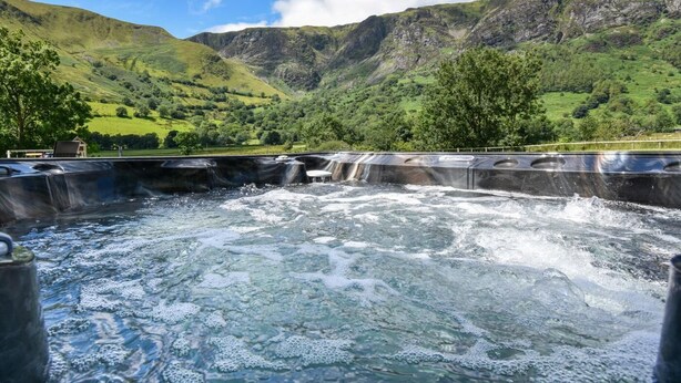 Eine heiße Quelle mit Wasserfall, umgeben von grünen Hügeln und Bergen.