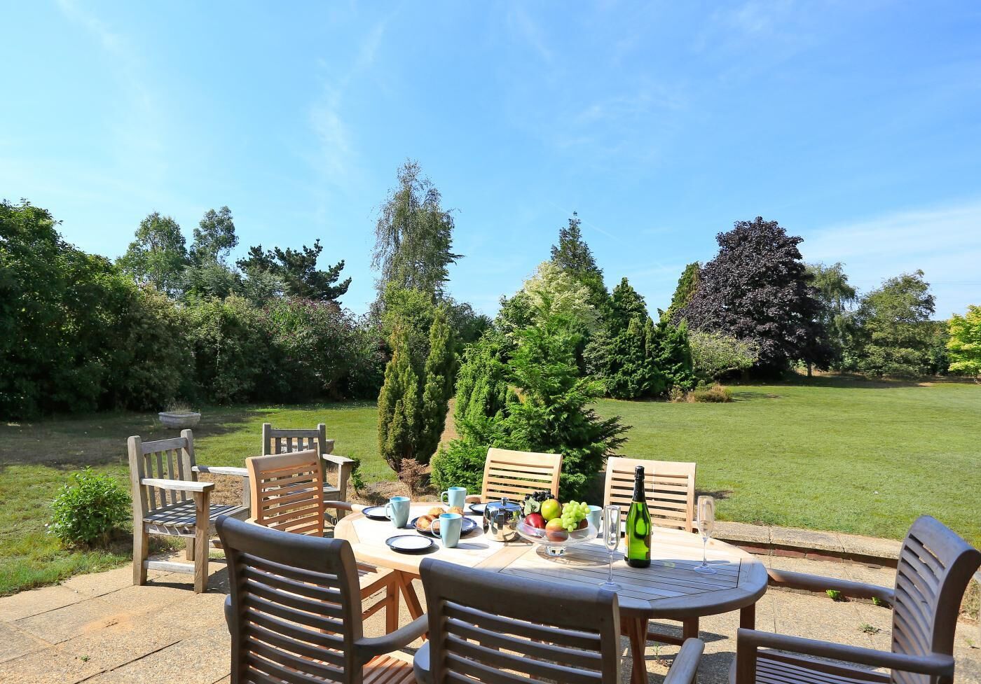 An outdoor dining area with a table set for four, surrounded by chairs, with a view of a well-maintained lawn and trees.