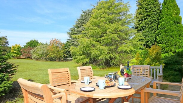 A wooden outdoor dining table with six chairs, surrounded by lush greenery and trees.