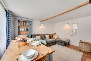 A dining area with a wooden table set for four, a bookshelf, a grey sofa with cushions, and a woven basket. - 2 Bed in Cley (oc-1776) (Cley next the Sea)