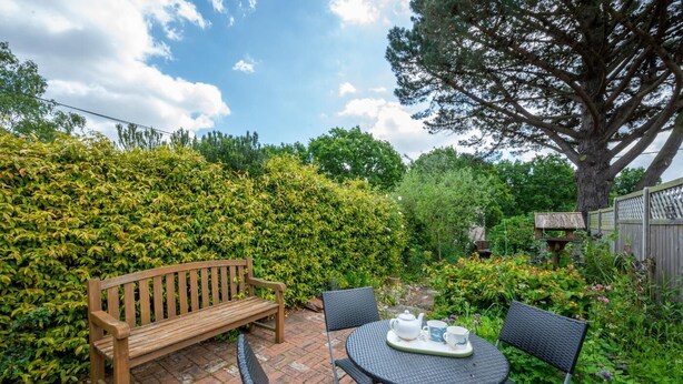 A patio with a wooden bench and a round table with chairs, surrounded by lush greenery and a large tree. - 2 Bed in Westleton (oc-gors) (Westleton)