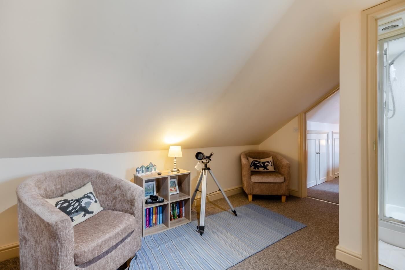 A cozy attic room with a chair, a small table with a lamp, a bookshelf, and a tripod.