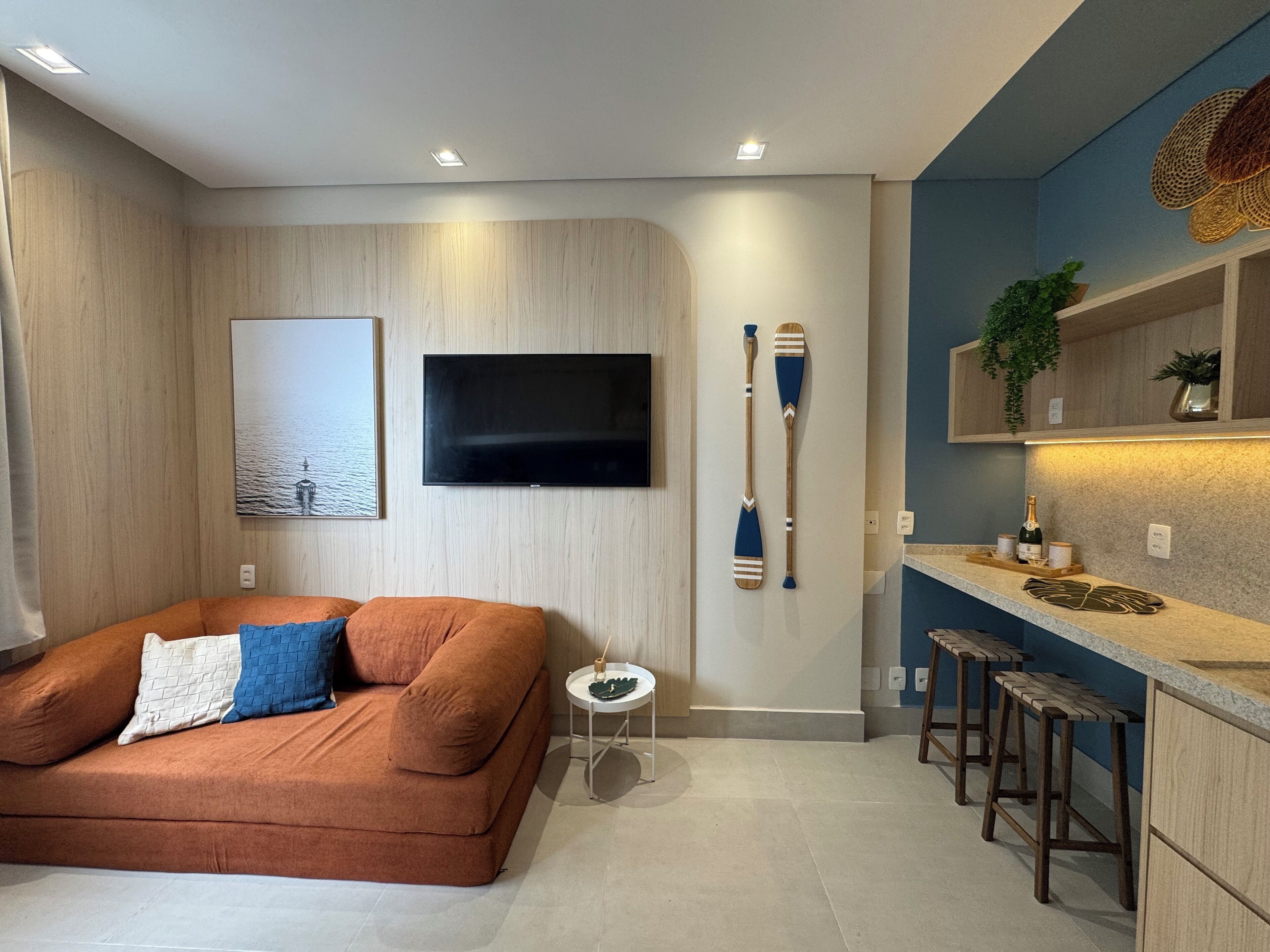 Un salon moderne comprenant un canapé, une petite table d’appoint, un téléviseur fixé au mur et un coin cuisine avec des tabourets de bar et un comptoir.
