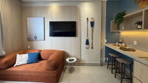 A modern living room with a sofa, a small side table, a TV mounted on the wall, and a kitchen area with bar stools and a countertop. - Sea View • 3 King Beds • Premium Comfort in BC (Balneário Camboriú)