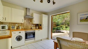 A compact kitchen with a washing machine, oven, and microwave. There is a dining area with a table and chairs, and a view of the garden outside. - 1 Bed in Stanhoe (oc-1792) (Stanhoe)