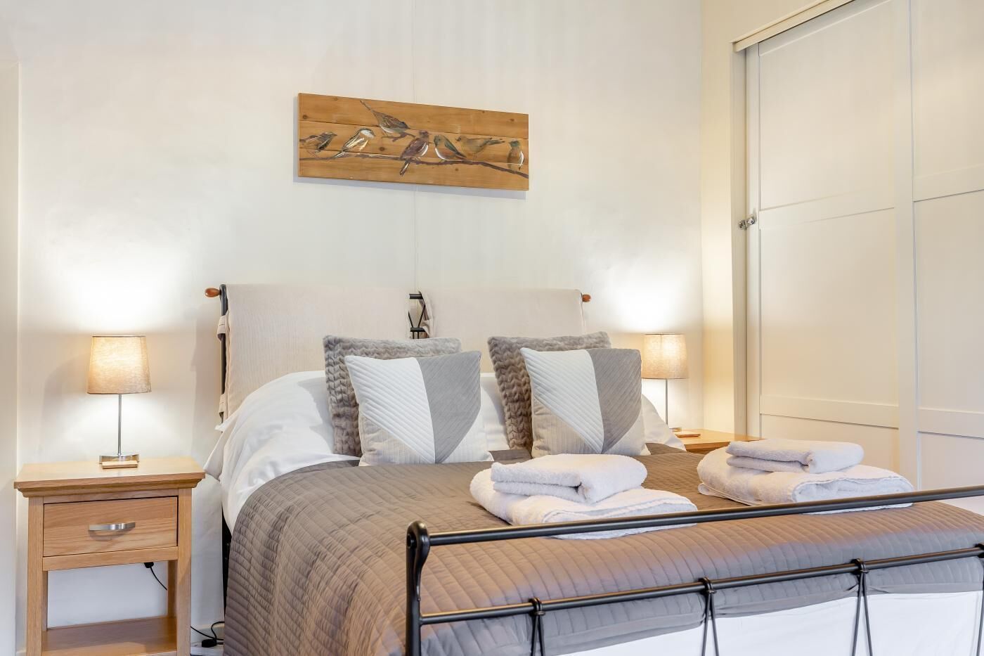 Een netjes opgemaakt bed met een hoofdbord, twee nachtkastjes met lampen, en een aan de muur hangend kunstwerk van vogels op een tak.