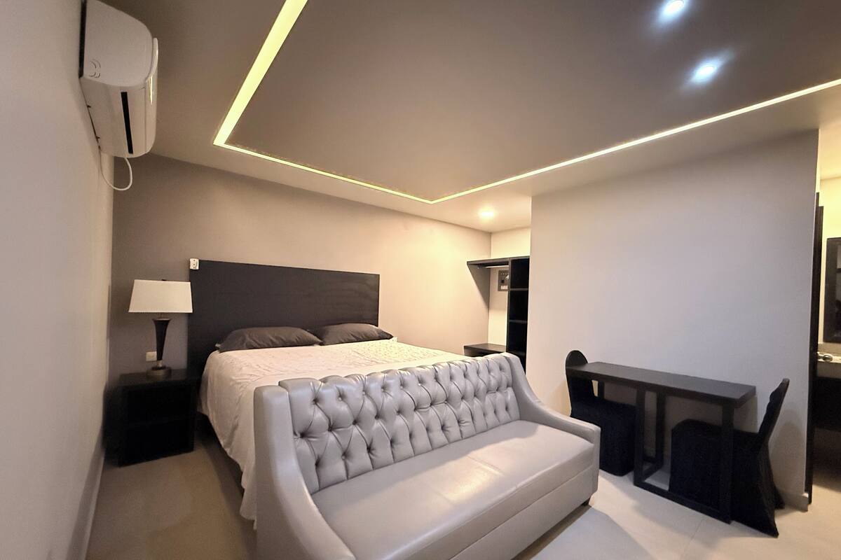 A modern bedroom with a large bed, a grey sofa, a desk, and a wardrobe.