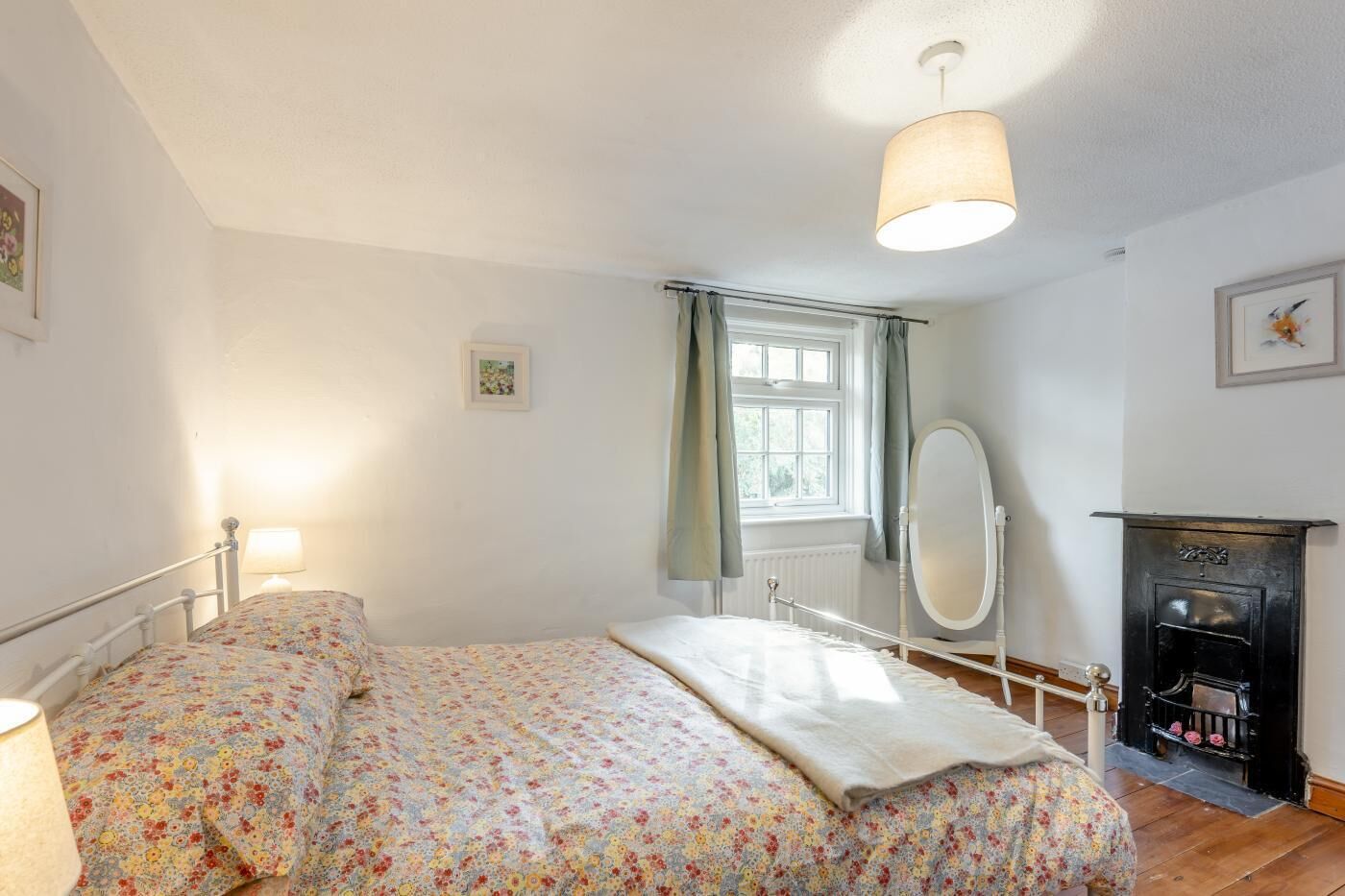 A bedroom with a bed, a bedside lamp, a window with curtains, a mirror, and a fireplace.