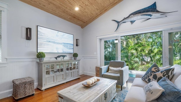 A spacious living room with a wooden ceiling, a large window with a view of greenery, a white sofa with patterned cushions, and a white cabinet with glass doors. - Spacious Beachfront Home in Holmes Beach on Anna Maria Island! Swell Life (Holmes Beach)