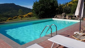 Ein Schwimmbecken mit klarem, blauem Wasser, umgeben von einer Ziegelterrasse, mit Blick auf eine gebirgige Landschaft.