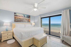Une chambre à coucher avec un lit, des lampes de chevet, un tableau, un ventilateur de plafond et une vue sur la plage, visible à travers une grande fenêtre.