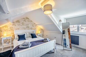 Une chambre avec un plafond en pente, un lit doté d’une tête de lit blanche, deux tables de chevet avec des lampes et un grand miroir.