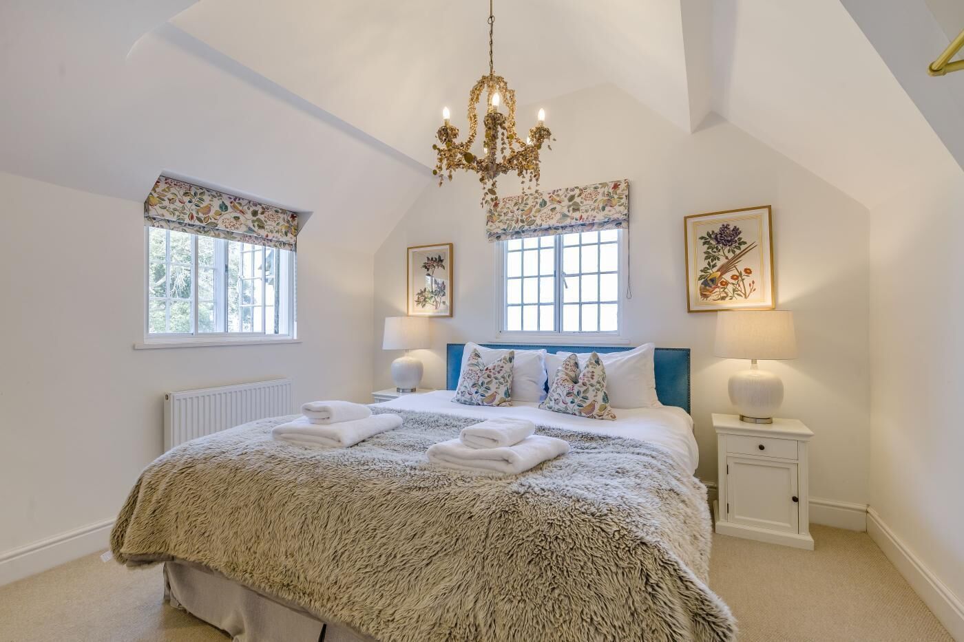 A bedroom with a large bed, two bedside tables, a chandelier, and two windows with curtains.