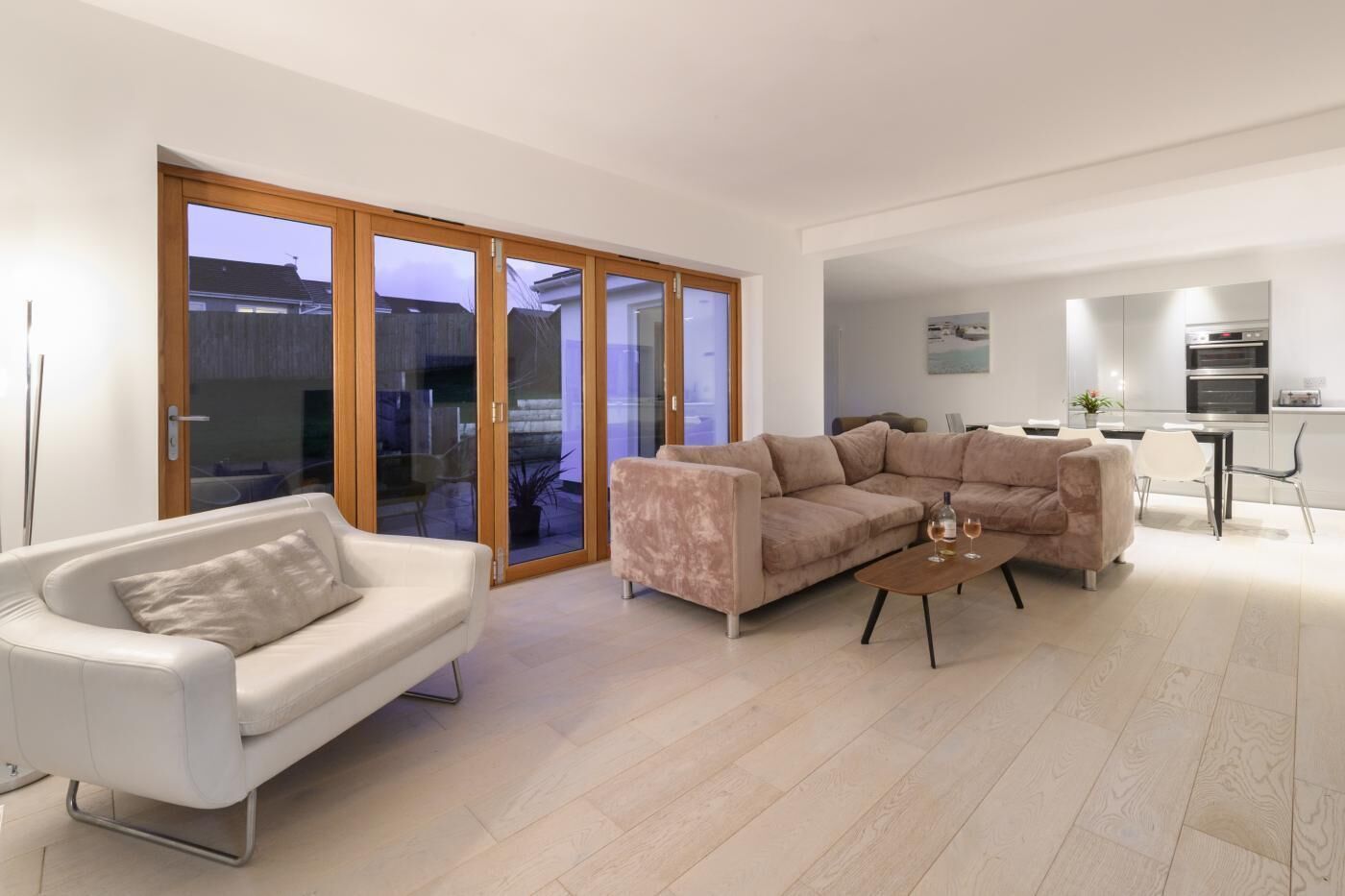 Un salon moderne avec un canapé blanc, un canapé d’angle beige et une table basse. Une porte-fenêtre coulissante donne accès à un espace extérieur.