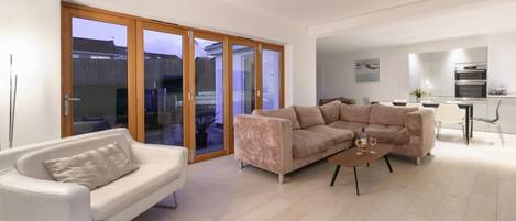 Un salon moderne avec un canapé blanc, un canapé d’angle beige et une table basse. Une porte-fenêtre coulissante donne accès à un espace extérieur.
