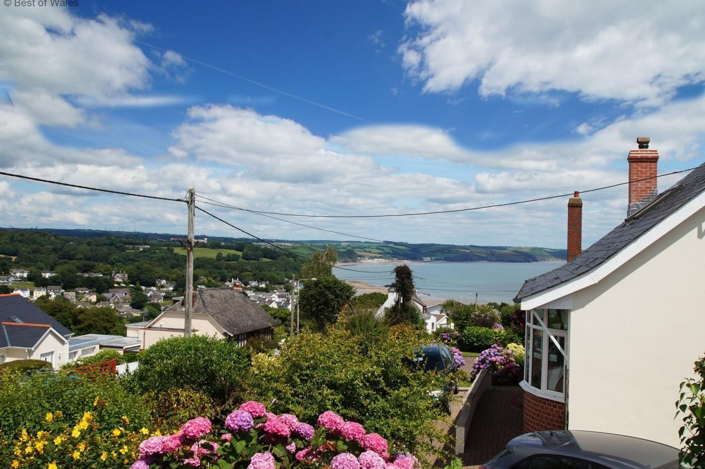 A coastal village with houses, a view of the sea, and a garden with pink flowers.