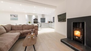 A modern living room with a sofa, a coffee table with glasses, a fireplace, and a kitchen in the background. - 4 Bed in Saundersfoot (oc-493314) (Saundersfoot)