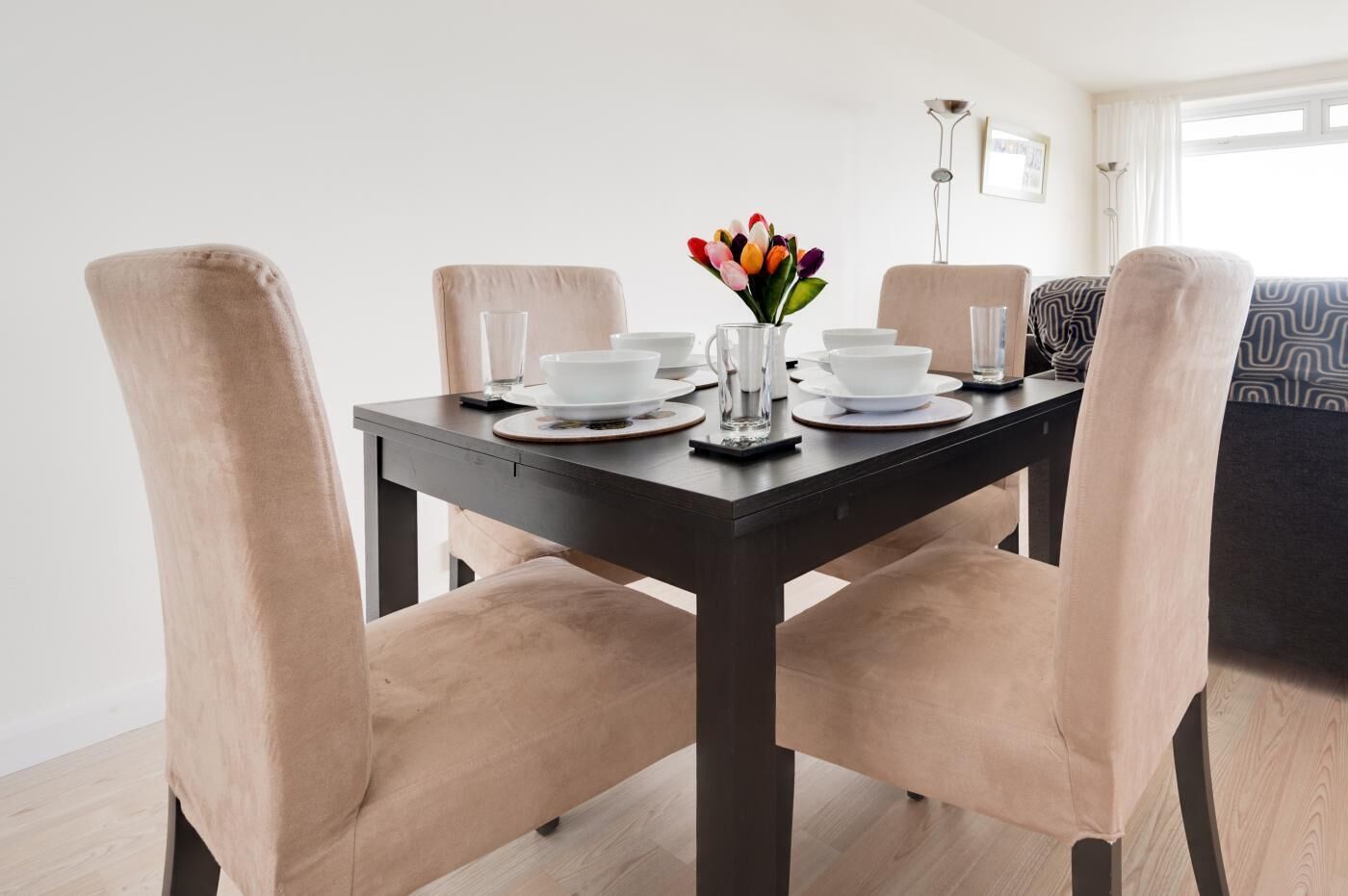A dining table with chairs, a vase of flowers, and a lamp in a room with a sofa and a window.