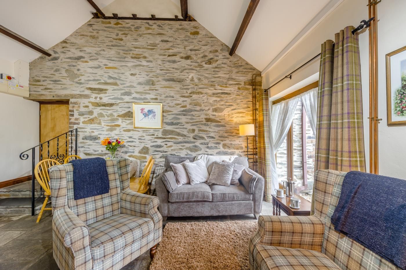 A cozy living room with a stone wall, a grey sofa, plaid armchairs, and a wooden staircase.