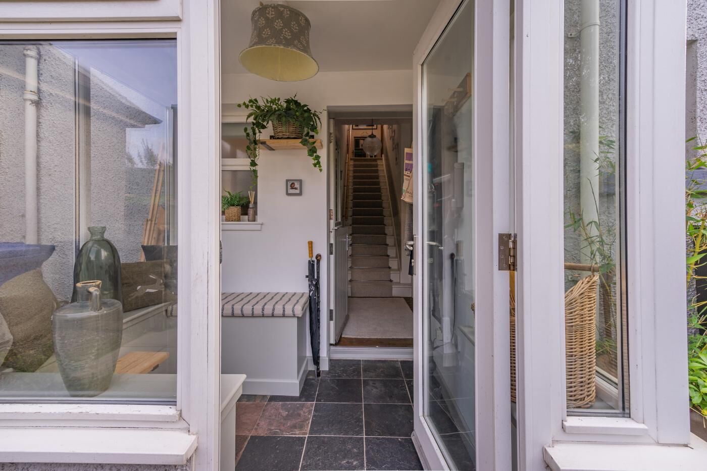 Une entrée moderne avec une porte en verre, un escalier menant à l’étage supérieur et un petit banc agrémenté d’un panier de plantes.