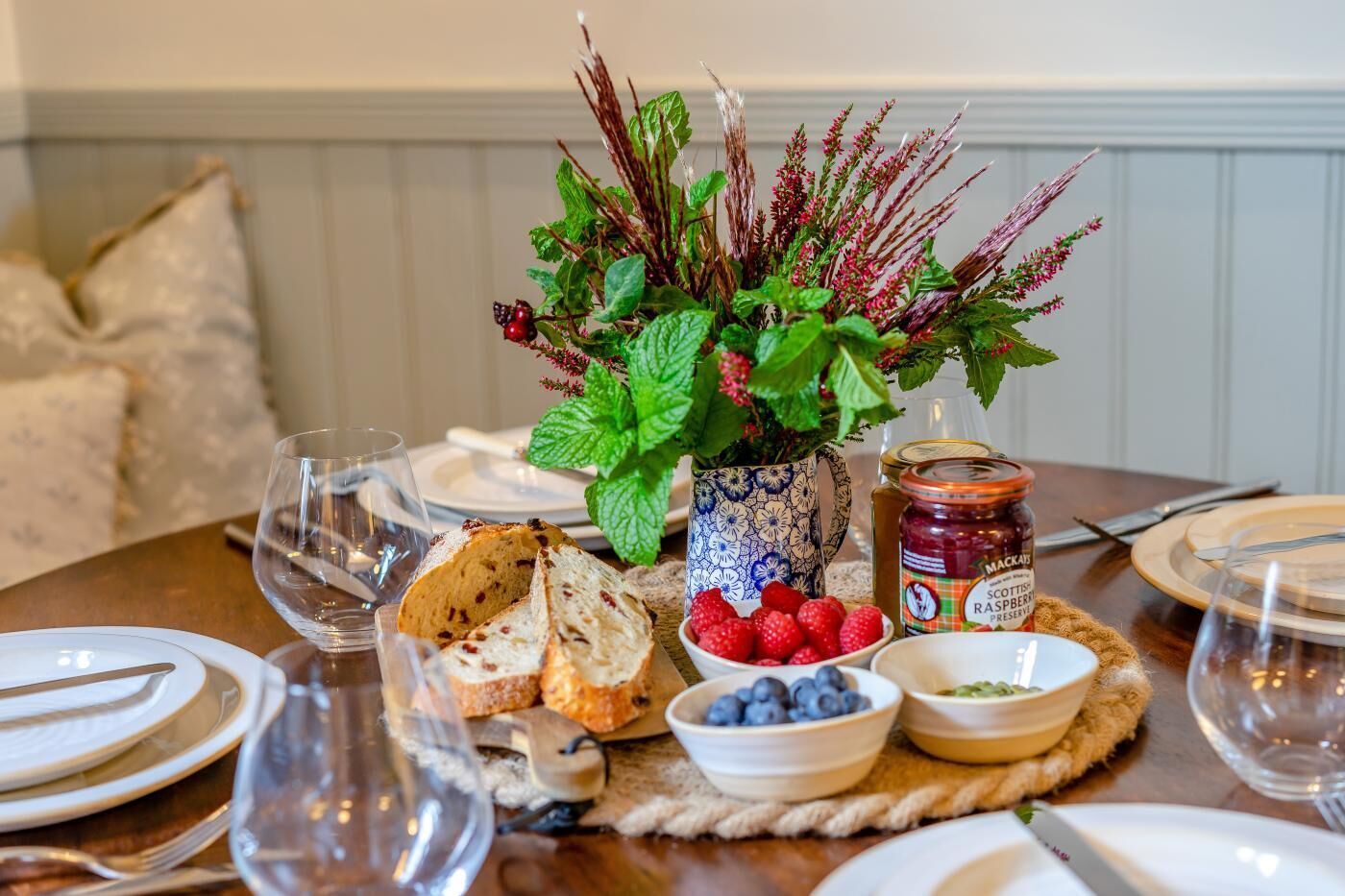 Une table à manger dressée avec des assiettes, des verres et un centre de table composé d’herbes fraîches et de baies.