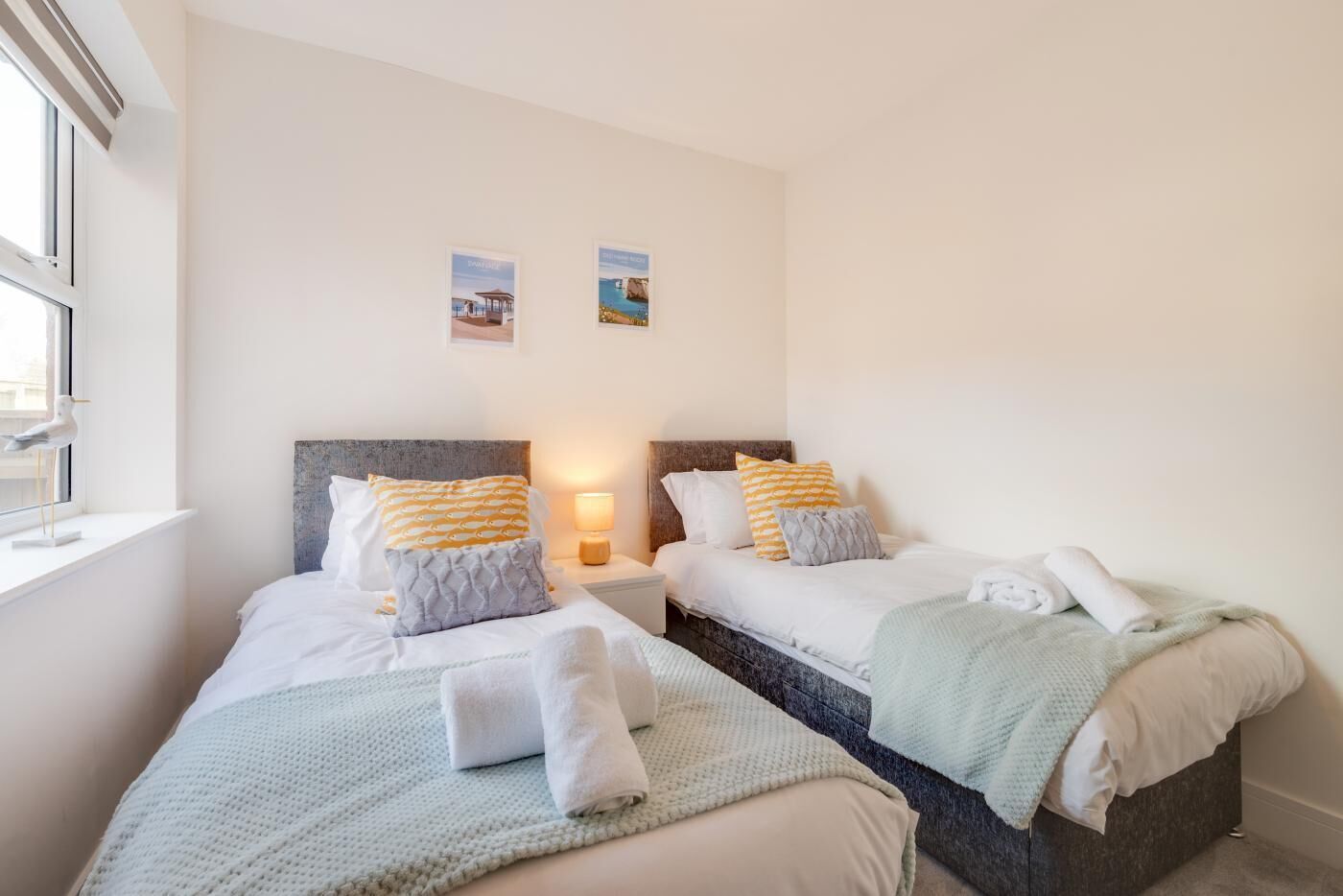 A double room with two beds, each with white bedding and decorative pillows. There is a window on the left side, and a small table with a lamp between the beds.